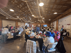 May show: People seated at round tables in a decorated hall, some wearing hats and colorful outfits, with string lights hanging from the ceiling.