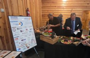 May show: People serve themselves food from a buffet table near a wooden wall, with a sponsor sign and certificates displayed on a separate table.